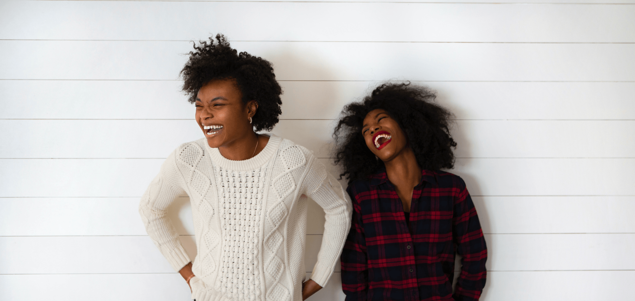 Two females laughing against a white wooden backdrop.