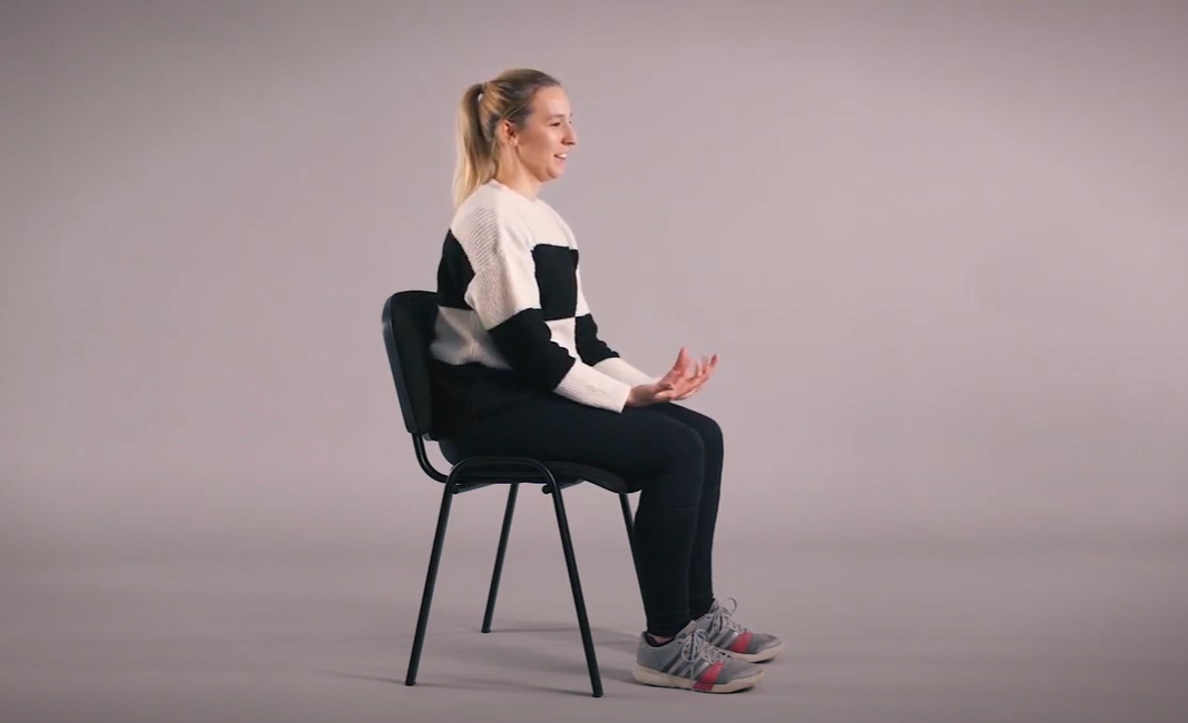 A female sitting on a chair in a room with a white backdrop talking about Chair Based Exercises.